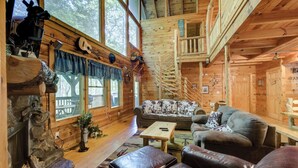L’intérieur d’une cabane en rondins est agrémenté d’une cheminée en pierre, de murs en bois, d’un escalier en colima çon et d’un coin salon confortable avec des canapés en cuir et une table basse.