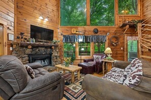 A log cabin interior with a stone fireplace, leather furniture, and large windows overlooking a forest. - Spacious Cabin w/ Pool Table, Hot Tub & Views (Sevierville)
