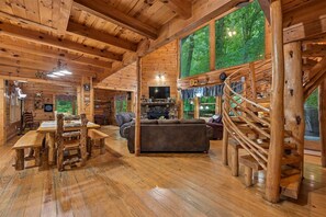 A wooden cabin interior with a dining area, sofa, TV, and a spiral staircase. - Spacious Cabin w/ Pool Table, Hot Tub & Views (Sevierville)