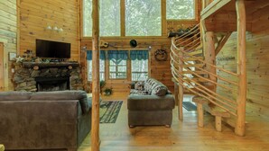 L’intérieur d’une cabane en rondins, avec une cheminée en pierre, un téléviseur, un canapé et un escalier en colimaçon.