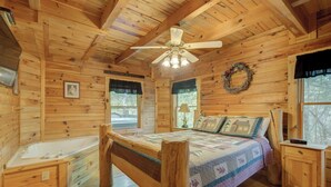 Une chambre dans une cabane en bois, avec un lit, une fenêtre, une couronne décorative accrochée au mur et un ventilateur de plafond.