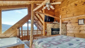 A log cabin room with a stone fireplace, a large window with a view of the mountains, a wooden ceiling, and a bed with a quilt. - Memorable Mountain Views w/ Hot Tub (Sevierville)