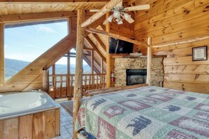 A log cabin room with a stone fireplace, a large window with a view of the mountains, a wooden ceiling, and a bed with a quilt.