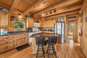 A wooden cabin interior with a kitchen featuring wooden cabinets, a central island with bar stools, a microwave, and a refrigerator.