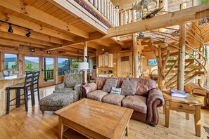 A wooden cabin interior with a sofa, chairs, a coffee table, a spiral staircase, and large windows offering a view of the outdoors. - Scenic Cabin w/ Hot Tub & Resort Amenities (Sevierville)