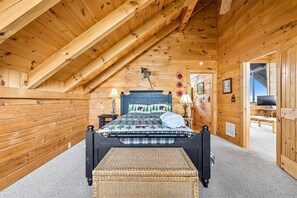 Een slaapkamer in een houten chalet, met een schuine plafond, een bed met een ruitdessin, twee bedlampen, een klein tafeltje en een televisie.