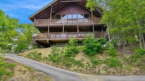 Een houten vakantiehuis met twee verdiepingen, inclusief een balkon op de bovenverdieping, omringd door bomen en een onverharde pad.