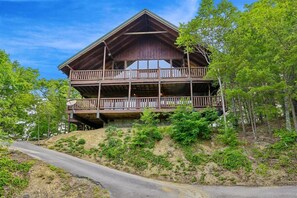A two-story wooden cabin with a balcony on the upper level, surrounded by trees and a dirt path. - Scenic Cabin w/ Hot Tub & Resort Amenities (Sevierville)