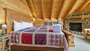 A wooden cabin bedroom with a stone fireplace, a quilted bedspread, and a nightstand with a lamp.