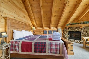 A wooden cabin bedroom with a stone fireplace, a quilted bedspread, and a nightstand with a lamp. - Scenic Cabin w/ Hot Tub & Resort Amenities (Sevierville)
