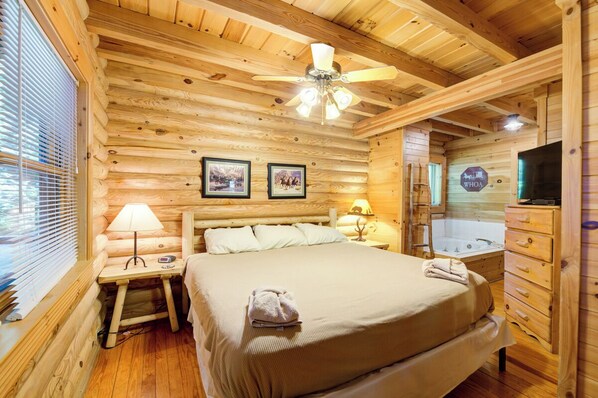Un dormitorio tipo cabaña de troncos con una cama, dos lámparas de noche, un ventilador de techo, una ventana con persianas, un tocador de madera y un televisor montado en la pared.