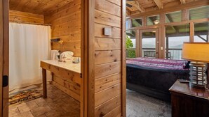 Une cabane en bois avec une salle de bain et une chambre. La salle de bain est équipée d’un rideau de douche, d’un porte-serviettes et d’un lavabo. La chambre comprend un lit, une lampe et une fenêtre donnant sur l’extérieur.