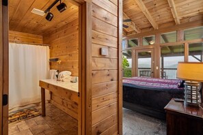 Une cabane en bois avec une salle de bain et une chambre. La salle de bain est équipée d’un rideau de douche, d’un porte-serviettes et d’un lavabo. La chambre comprend un lit, une lampe et une fenêtre donnant sur l’extérieur.