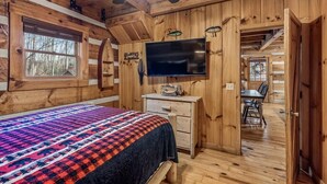 Ein Schlafzimmer in einer Blockhütte mit Bett, Kommode, Fernseher und einem Essbereich, der durch eine offene Tür sichtbar ist.
