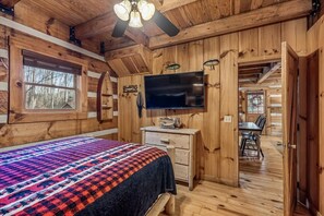 A log cabin bedroom with a bed, dresser, TV, and a dining area visible through an open door.