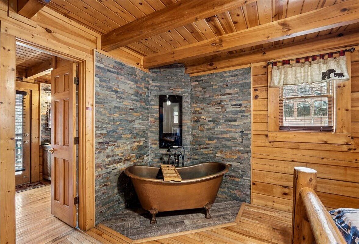 A rustic bathroom with a freestanding bathtub, a stone wall, and wooden beams.