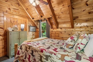Un dormitorio tipo cabaña de madera con una cama, una mesita de noche, una ventana y un ventilador de techo.