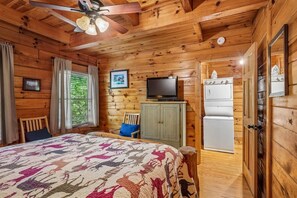 A wooden cabin room with a bed, a television, a refrigerator, and a ceiling fan.
