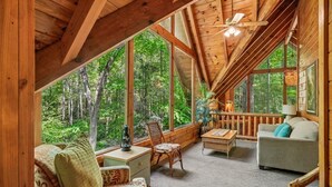 Ein gemütliches Blockhaus-Interieur mit einem großen Fenster mit Blick auf einen Wald, einem Deckenventilator und einer Sitzecke mit einem Tisch.