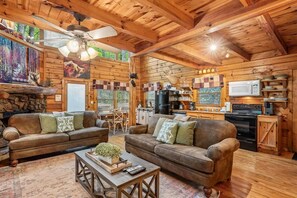 A log cabin interior with two sofas, a coffee table, a ceiling fan, and a kitchen area with a microwave.