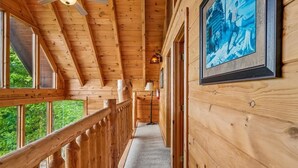 Un couloir intérieur en bois, avec un ventilateur de plafond, un tableau encadré au mur et une vue sur la verdure à l’extérieur.