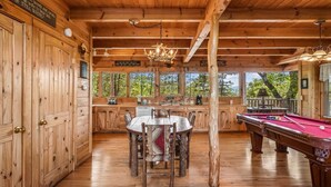 L’intérieur d’un chalet en bois comprend une table à manger, des chaises, une table de billard et une grande fenêtre donnant sur un paysage verdoyant.
