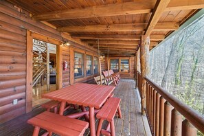A wooden cabin with a covered porch, picnic tables, and a view of the forest.