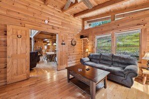 A wooden interior with a sofa and a coffee table, a dining area with chairs, and a view through large windows.