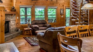 A cozy cabin interior with a stone fireplace, leather sofa, wooden dining table, and large windows offering a view of the outdoors. - Rustic Wooded Cabin w/ Game Room & Hot Tub (Sevierville)