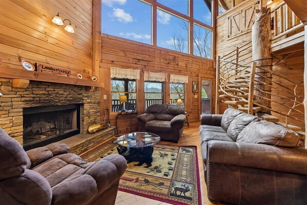 A log cabin interior with a stone fireplace, leather sofas, and a large window overlooking a scenic landscape.