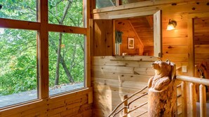 A wooden cabin with a large window overlooking a forest, a wooden bear sculpture, and a staircase. - Rustic Wooded Cabin w/ Game Room & Hot Tub (Sevierville)
