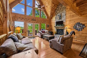 A log cabin interior with a fireplace, wooden walls, and a large window overlooking greenery. - Rustic Luxe Charm w/ Views, Fireplace & Hot Tub (Sevierville)