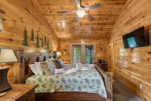 A wooden cabin bedroom with a bed, a ceiling fan, a television, and a window with blinds. - Rustic Luxe Charm w/ Views, Fireplace & Hot Tub (Sevierville)