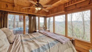 Une chambre à coucher située dans une cabane en bois, avec un lit, un ventilateur de plafond, de grandes fenêtres et une vue sur les arbres.