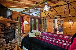 A wooden cabin interior with a stone fireplace, a bed with a red and black checkered blanket, and a television on the left wall.