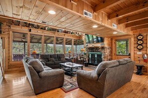 A cozy log cabin interior with a fireplace, a sofa set, and a large window overlooking greenery.