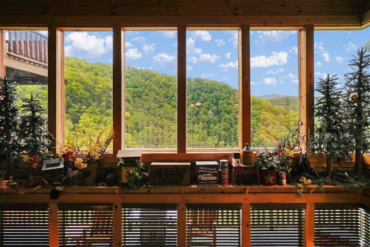 A room with large windows overlooking a forested hillside, wooden beams, and a shelf with various items including a sign that reads 'Country Living.'