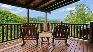 Una terraza de madera con dos mecedoras y una mesita, con vista a un paisaje boscoso.