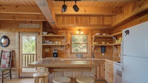A wooden cabin interior with a kitchen area featuring a central island, stools, a refrigerator, and shelves with dishes.