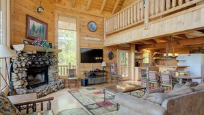 A rustic cabin interior with a stone fireplace, wooden furniture, a TV, and a balcony.