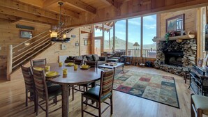 A wooden cabin interior with a dining area, a stone fireplace, and a large window overlooking a mountainous landscape.