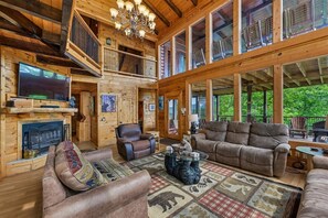 A spacious log cabin interior with a fireplace, comfortable seating, and large windows offering a view of the outdoors. - 7-Bedroom Cabin with Game Room & Hot Tub (Sevierville)