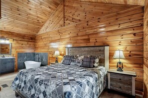 A wooden cabin bedroom with a bed, bedside lamps, a sink, and a bathtub.