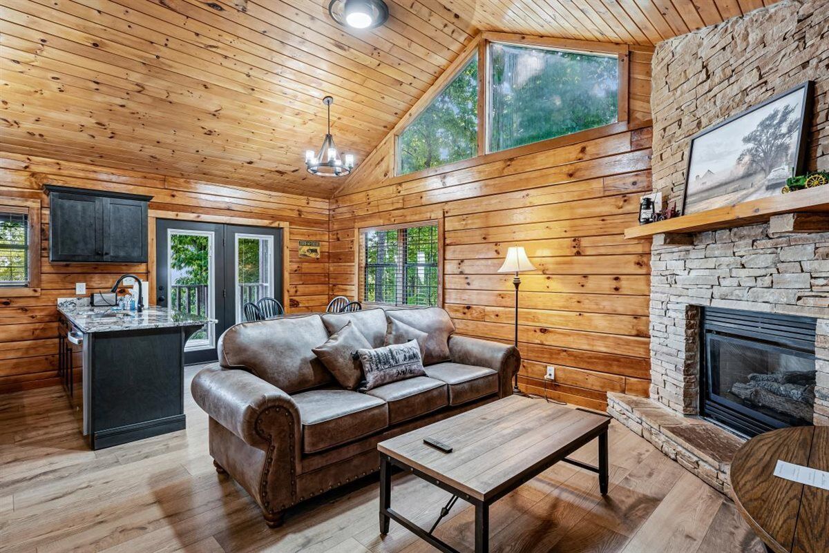 A log cabin interior with a wooden ceiling, a stone fireplace, a leather sofa, a wooden coffee table, and a kitchen area.