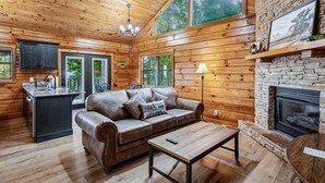 Ein Blockhaushaus-Interieur mit Holzbalkendecke, Kamin aus Stein, einer Ledersofa, einem Holztisch und einer Küchenzeile.