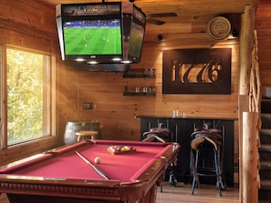 A wooden room with a pool table, a television displaying a sports game, and a fireplace.