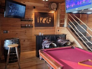 Una mesa de billar con superficie de fieltro rojo, paredes de madera, un televisor montado y una zona de bar con taburetes y un barril.