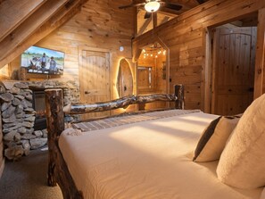A cozy log cabin bedroom with a stone fireplace, a wooden bed, and a television mounted on the wall.