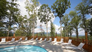 Zona de piscina con sillas de descanso y una terraza de madera con vistas a un paisaje boscoso.
