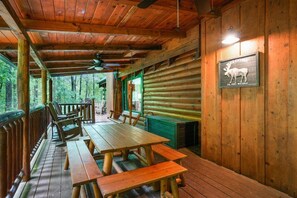A wooden cabin interior with a dining area, benches, a table, and a sign reading 'WELCOME TO THE WOODS'.